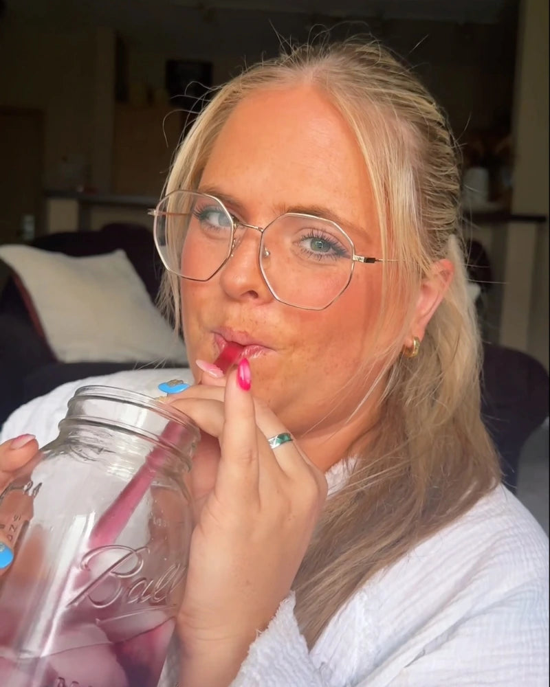 A person with long blonde hair and round glasses sips from a jar using a straw. They are wearing a white top and have colorful nails. The background shows a couch with a pillow.